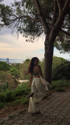 a woman walking down a path next to a tree and grass covered hillside with the ocean in the background