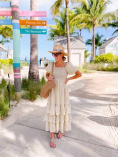 a woman in a white dress and hat standing on the side of a road with palm trees