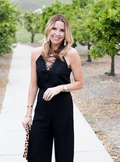 a woman in black jumpsuits and heels standing on the sidewalk with trees behind her