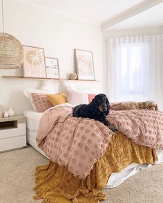 a black dog laying on top of a bed covered in pink and gold blankets next to a window