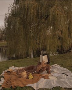 a woman reading a book while laying on a blanket in the grass near a tree