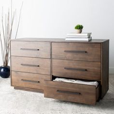 a wooden dresser with two drawers and a plant on top, in front of a white wall