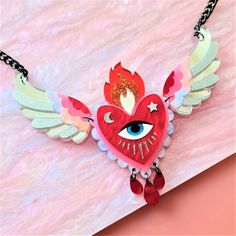 a red heart with wings and an evil eye on it's face is hanging from a black chain