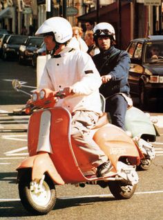 two people riding on the back of a red scooter down a city street