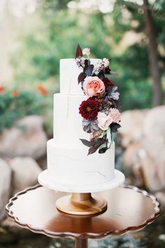 a white wedding cake with flowers on top sitting on a gold stand in front of some rocks