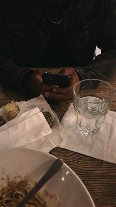 a man sitting at a table looking at his cell phone and eating pasta with a glass of water