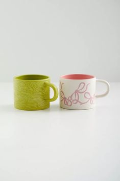 two coffee mugs sitting next to each other on a white counter top, one with pink and green designs