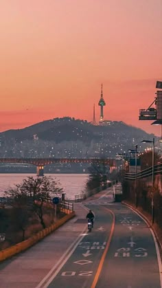 a person riding a bike down a street next to the ocean at sunset with a city in the background