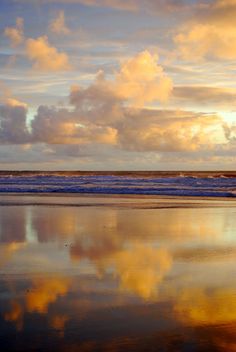 two people walking on the beach with their surfboards in hand and clouds reflected in the wet sand