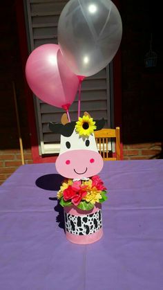 a cow with flowers and balloons sitting on top of a purple tablecloth covered table