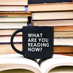 a black coffee mug that says what are you reading now? on top of a pile of books