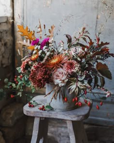 a vase filled with lots of flowers sitting on top of a wooden stool next to a wall