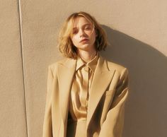 a woman leaning against a wall wearing a tan suit and gold shirt with her hands in her pockets