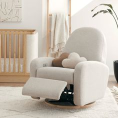 a rocking chair in a white room with a baby crib and potted plant