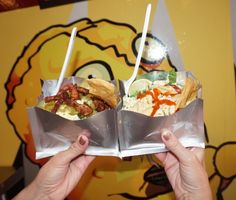 two burritos are being held up in front of a wall with colorful art