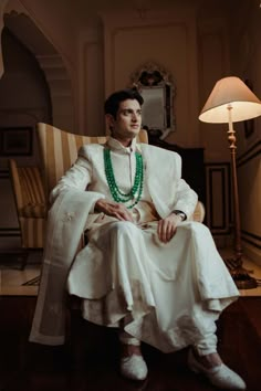 a man sitting in a chair with a green beaded necklace on his neck and wearing a white suit