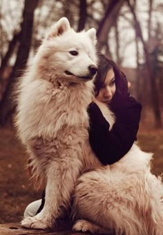 a woman hugging a large white dog on top of a wooden bench in the woods