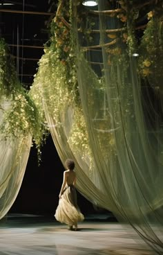 a woman is walking down the runway in front of sheer curtains with flowers hanging from them