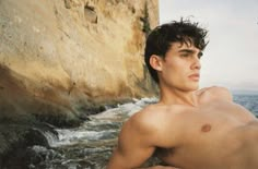 a shirtless young man sitting on the rocks by the ocean looking at the camera