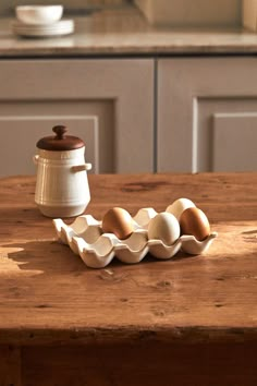 an egg tray with six eggs in it on a wooden table next to a coffee pot