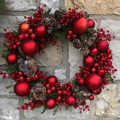 a christmas wreath with red ornaments and pine cones on a stone wall next to a brick wall