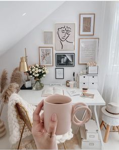 a person holding a pink coffee mug in their hand while sitting at a desk with pictures on the wall