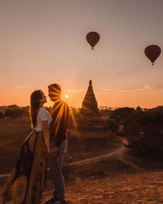 a man and woman standing in front of hot air balloons with the sun setting behind them