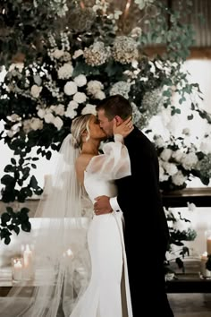 a bride and groom kissing in front of a floral backdrop at their wedding ceremony with candles