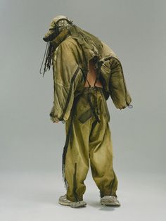 a man with dreadlocks standing in front of a gray background wearing a green outfit