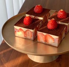 four pieces of cake with strawberries on top sitting on a gold plate next to a window