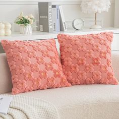 two pink pillows sitting on top of a white couch next to a book shelf and lamp