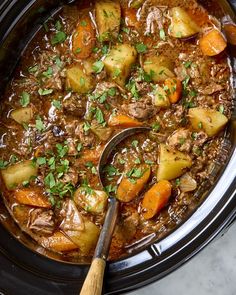 a crock pot filled with beef stew and carrots next to a wooden spoon