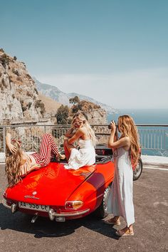 three women sitting on the back of an orange sports car in front of some mountains