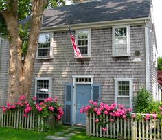 a gray house with pink flowers in the front yard and an american flag on the door