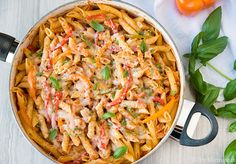 a pot filled with pasta and vegetables on top of a wooden table next to basil leaves