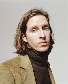 a man with long hair wearing a brown jacket and turtle neck sweater looking at the camera