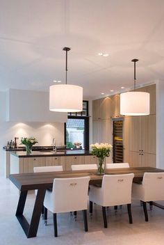 a dining room table with white chairs and lights hanging from it's ceiling in front of an open kitchen