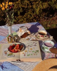 an open book on a picnic table with flowers and fruit in the middle, next to a bowl of strawberries