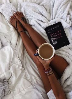 a woman laying in bed with her legs crossed holding a cup of coffee and book