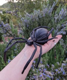 a person's hand holding a large black spider in front of purple wildflowers