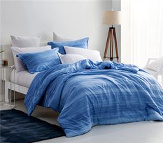 a bed with blue comforter and pillows in a white room next to a window