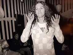 a man with long hair standing in front of a wall holding his hands up to the side