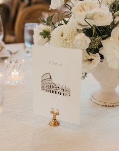 there is a card on the table with flowers in front of it and a vase full of white roses