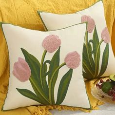 two pillows with pink flowers on them sitting on a yellow bed spread next to a bowl of fruit