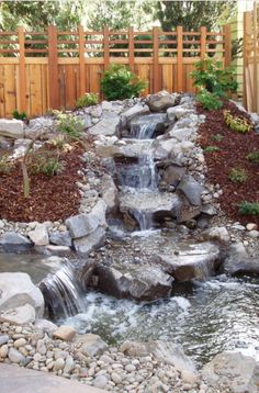 a small waterfall in the middle of a garden with rocks and gravel around it, surrounded by wood fence