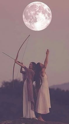 two women standing on top of a hill holding bows and arrows in front of the moon