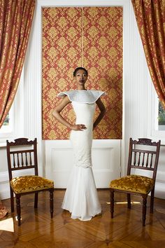 a woman in a white dress standing next to two chairs and a wall with red damask