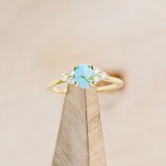 a gold ring with a turquoise stone surrounded by white diamonds on top of a wooden stand