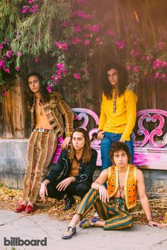 four young men sitting on a bench in front of pink and purple flowers, with one man looking at the camera