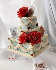 a three tiered cake decorated with red flowers and playing cards on the table next to it